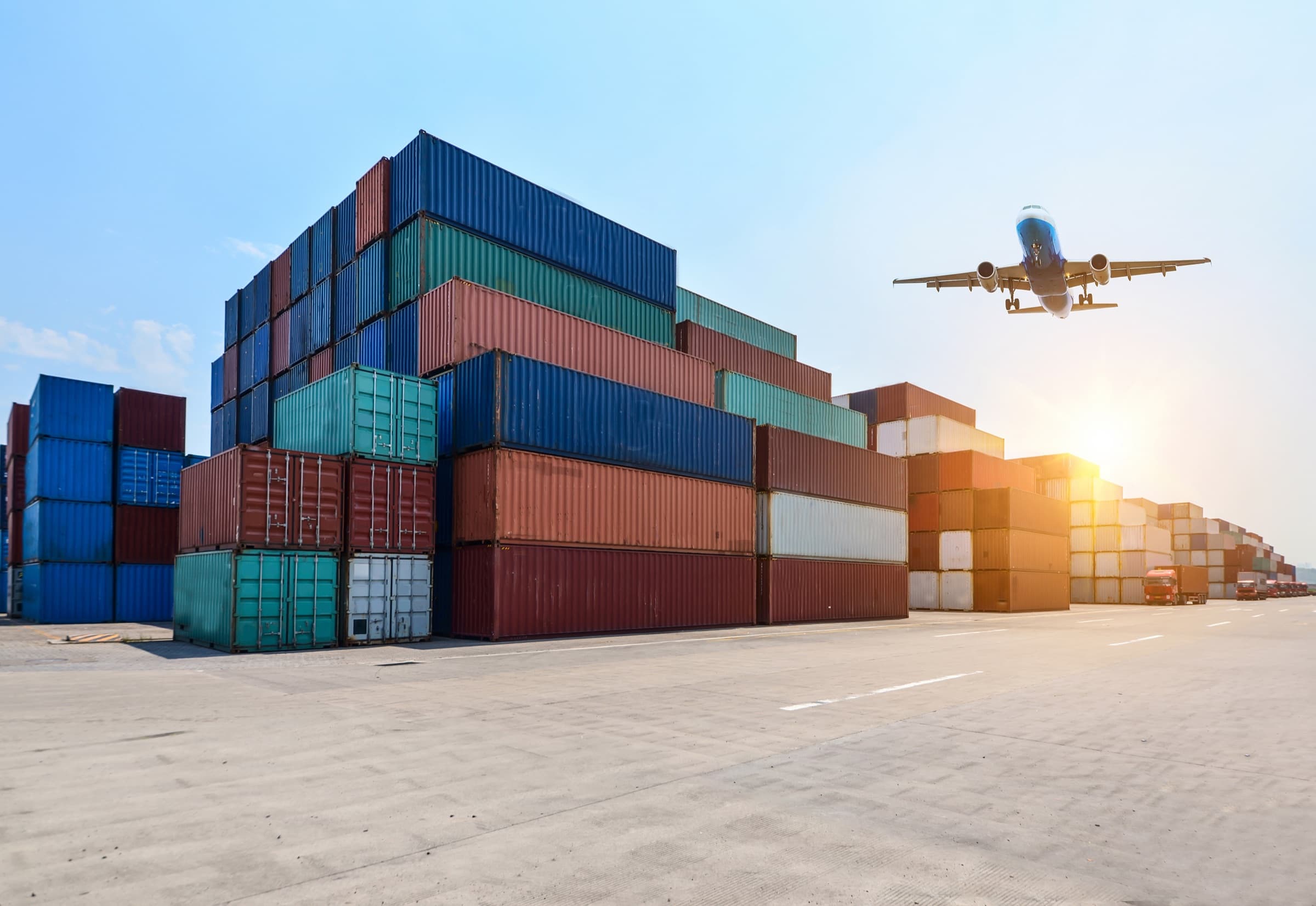 Shipping containers stacked at a busy port with a cargo plane departing overhead